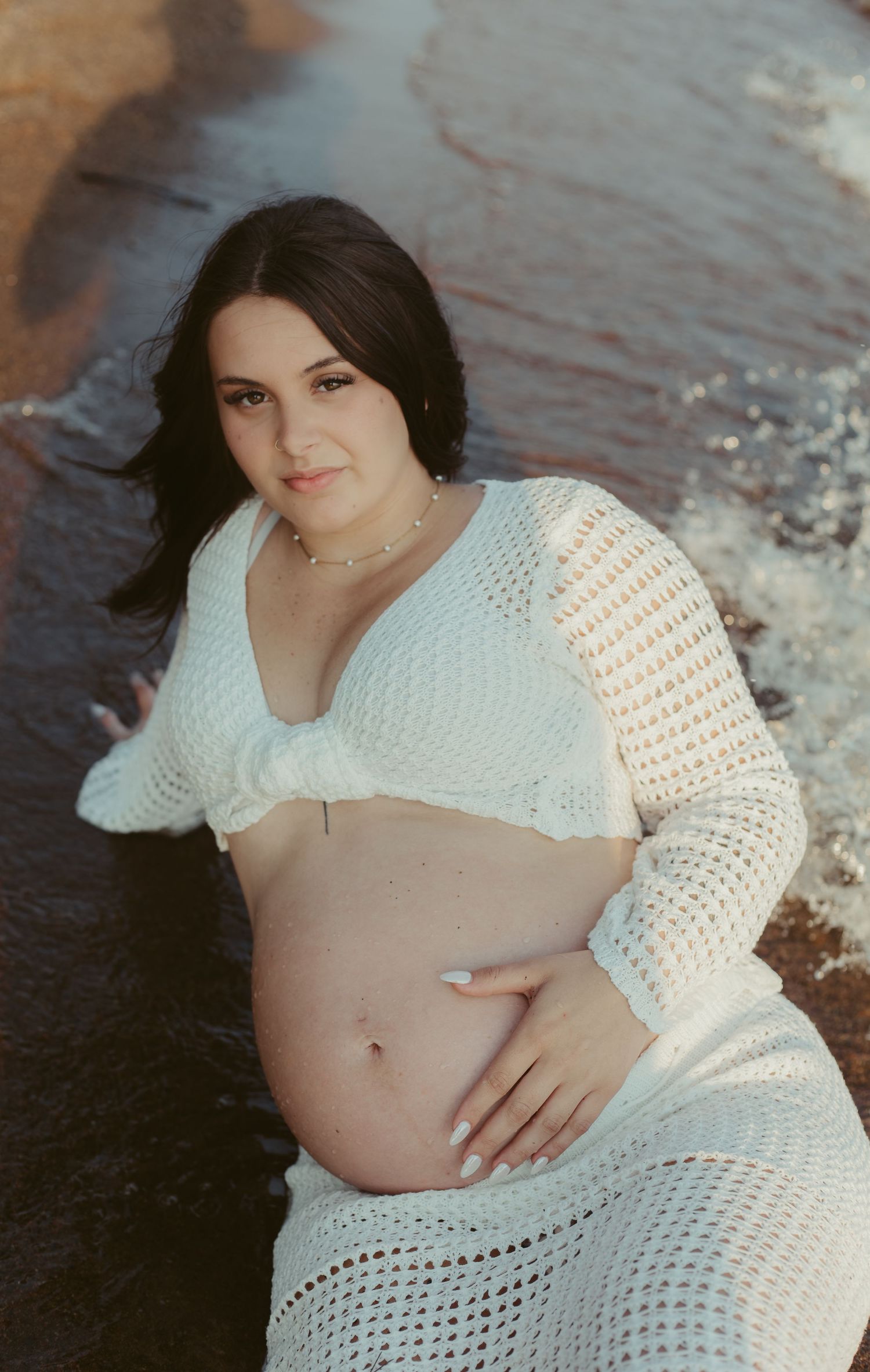 Pregnant woman in white crochet bikini top and skirt posing on beach, holding her baby bump.