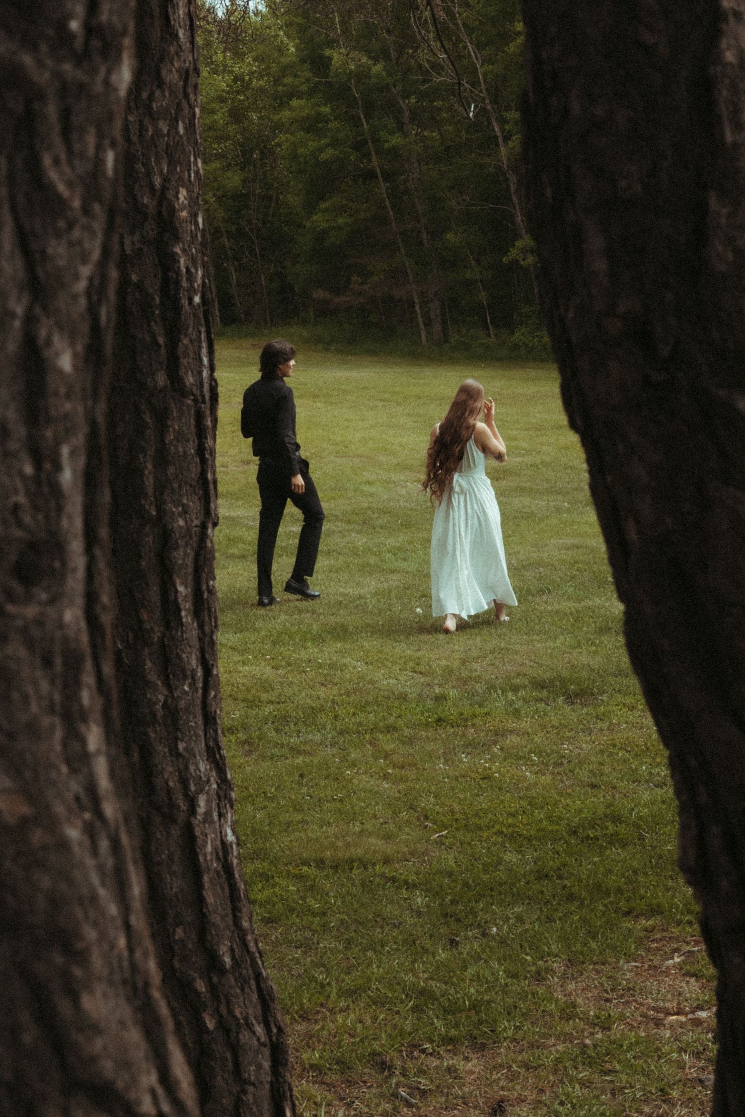 Couple in formal attire walking toward each other in a forest clearing between trees.