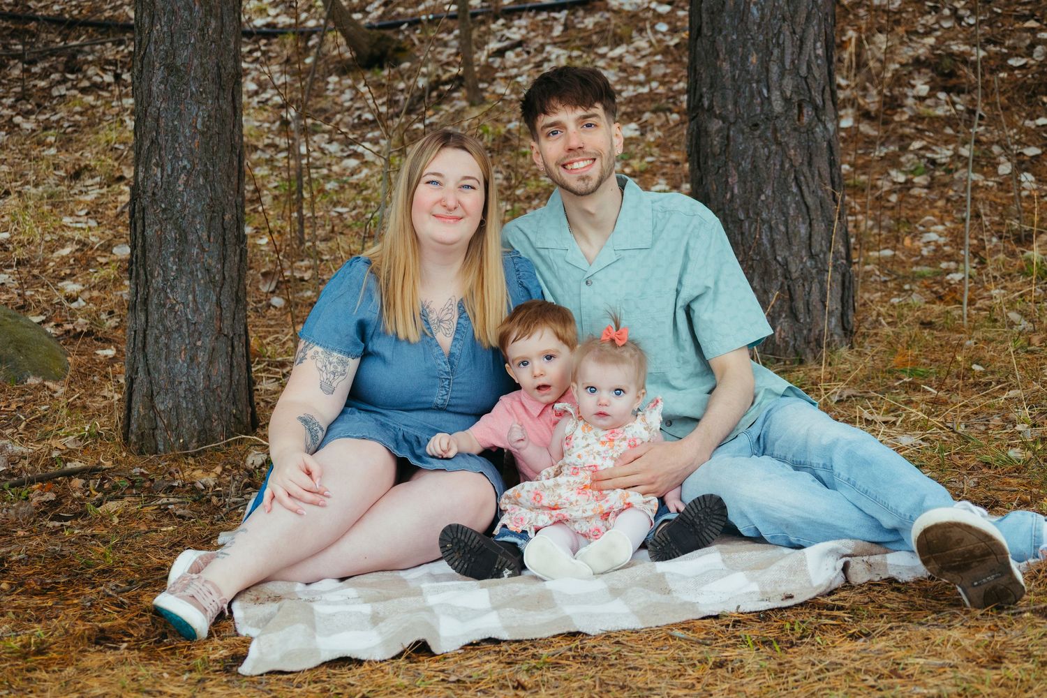 Young couple sits on blanket with two baby girls among pine trees in fall.