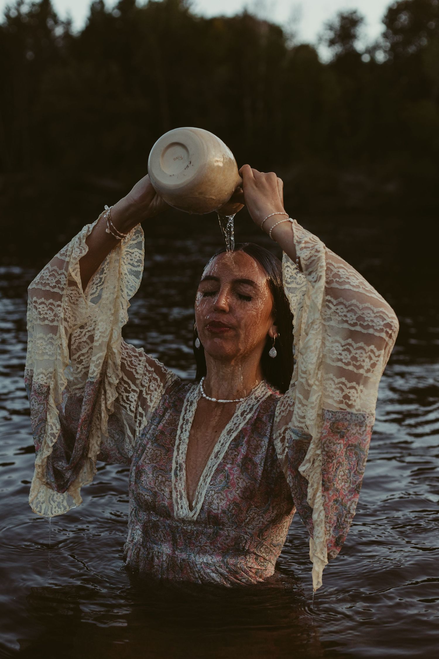 Woman in lace floral dress pouring water from ceramic jug over her head while standing in lake.