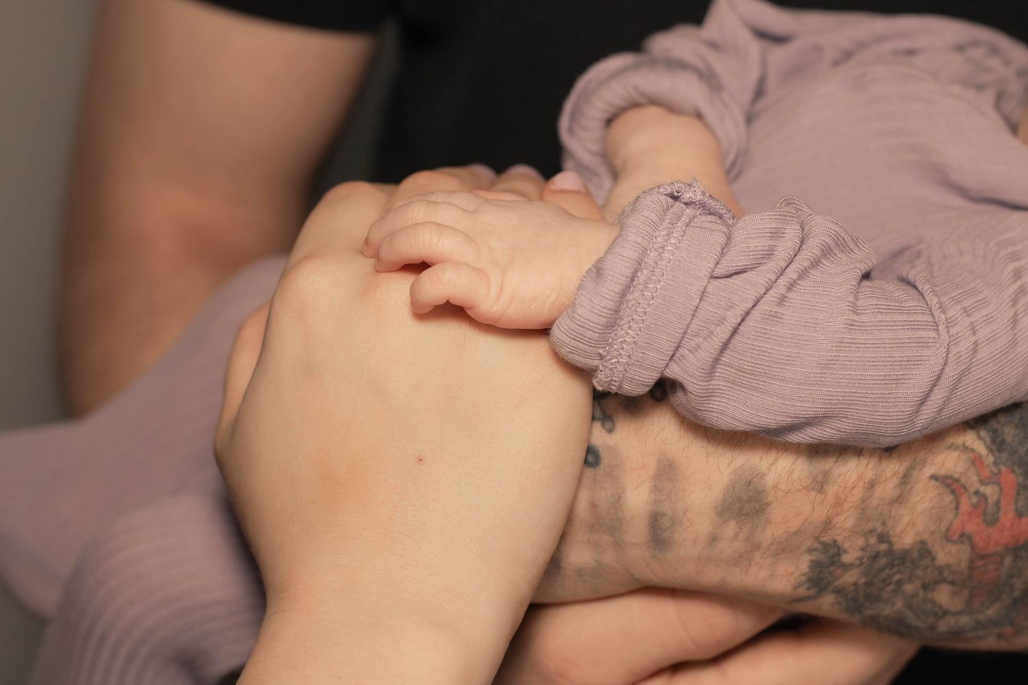 Tattooed arm gently holding a baby's tiny hand, close-up shot showing tender connection.