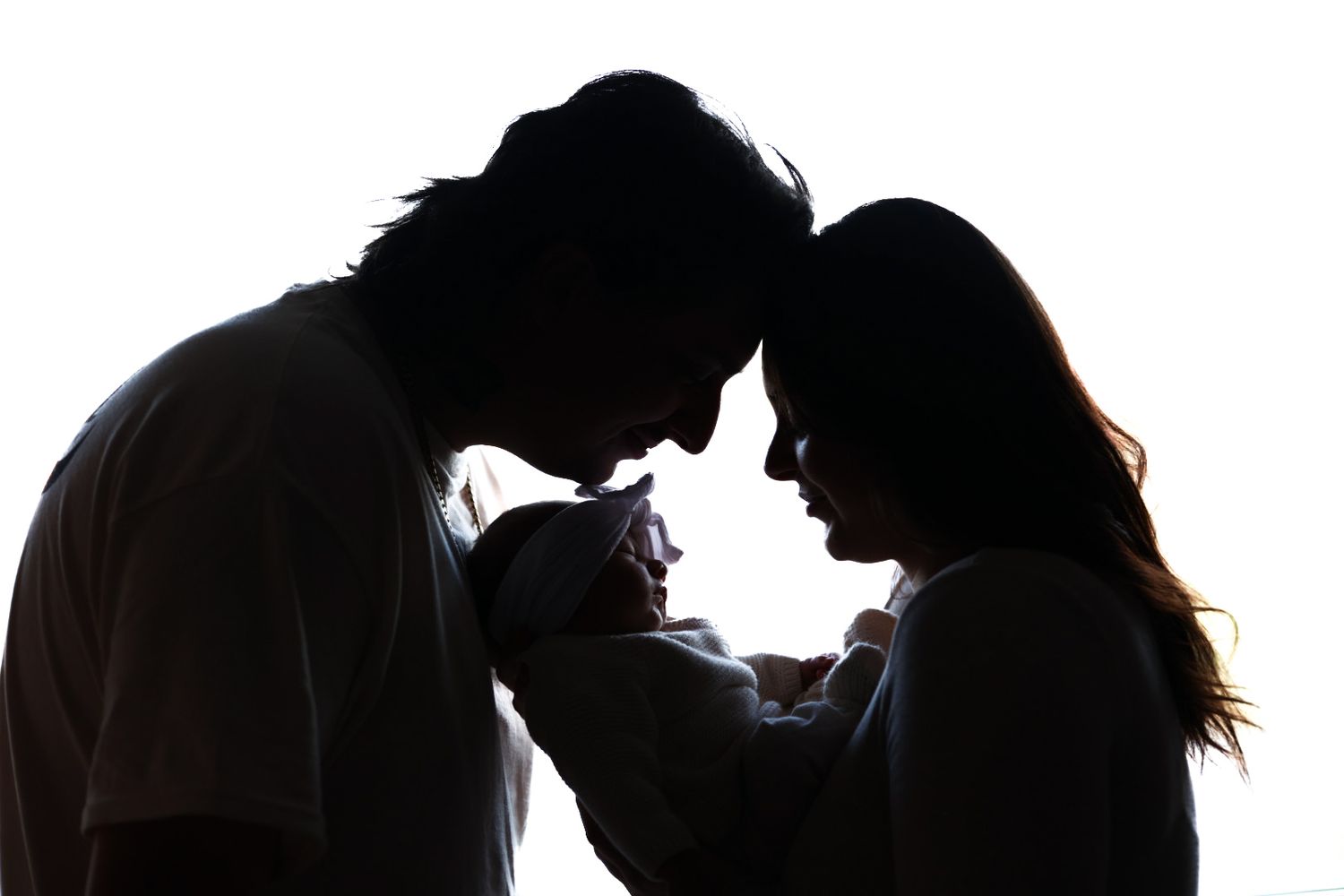 Silhouette of parents touching foreheads while holding their newborn baby against bright white background.