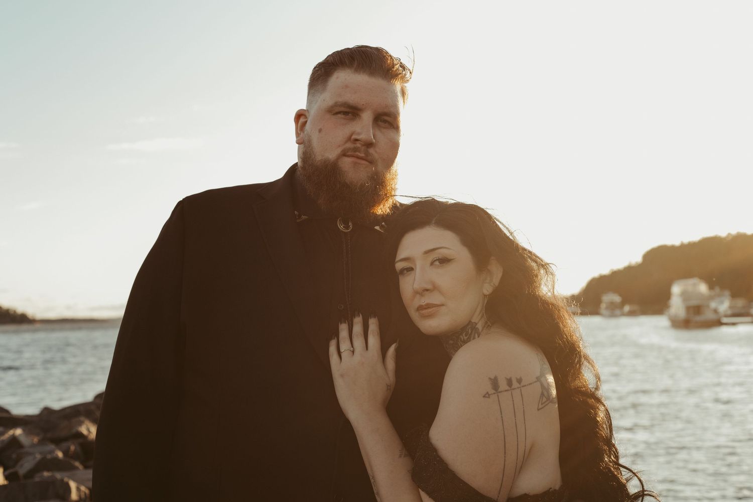 Couple standing together by the water at sunset, woman in black gown with tattoos.