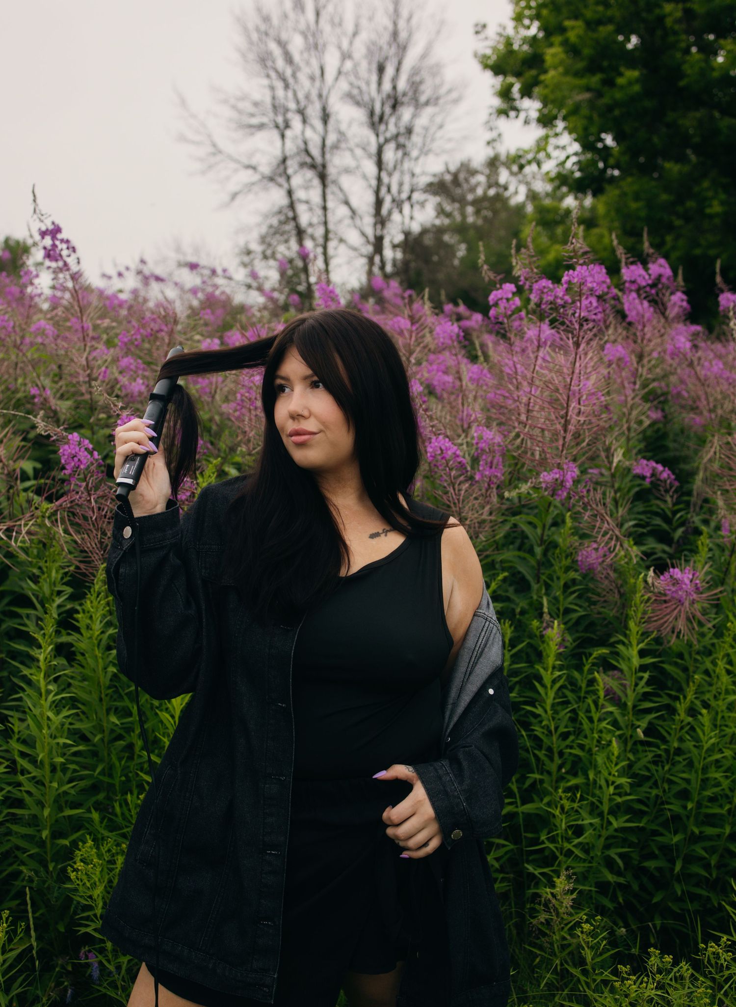 Woman in black outfit standing among vibrant purple wildflowers, holding her dark hair, with bare trees in background.
