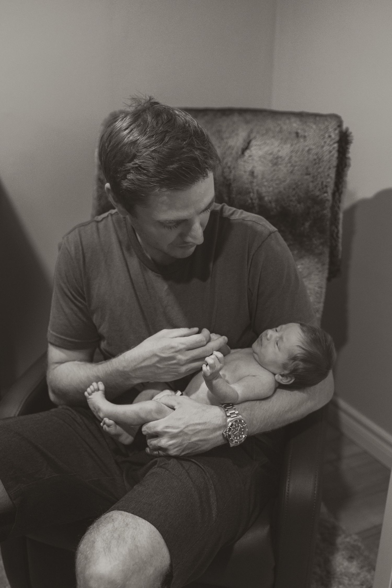 Black and white photo of a man sitting in a chair tenderly cradling a newborn baby in his arms.
