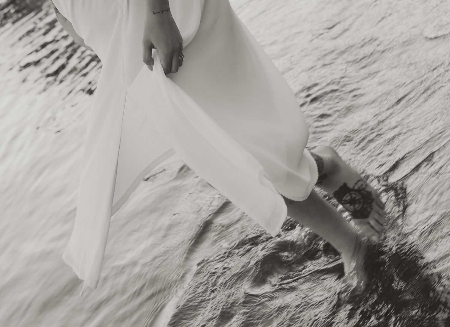 Black and white photo of a woman in a white dress standing at the edge of water, her bare feet touching the waves.