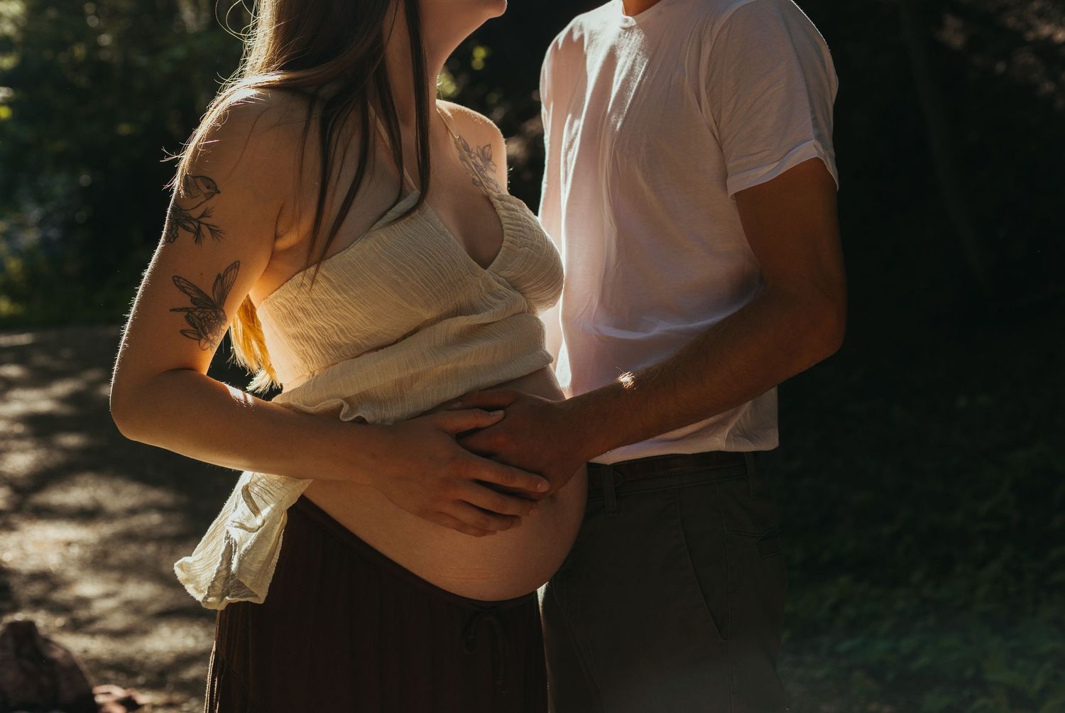 Couple embracing outdoors in dappled sunlight, partner holding pregnant woman's baby bump.
