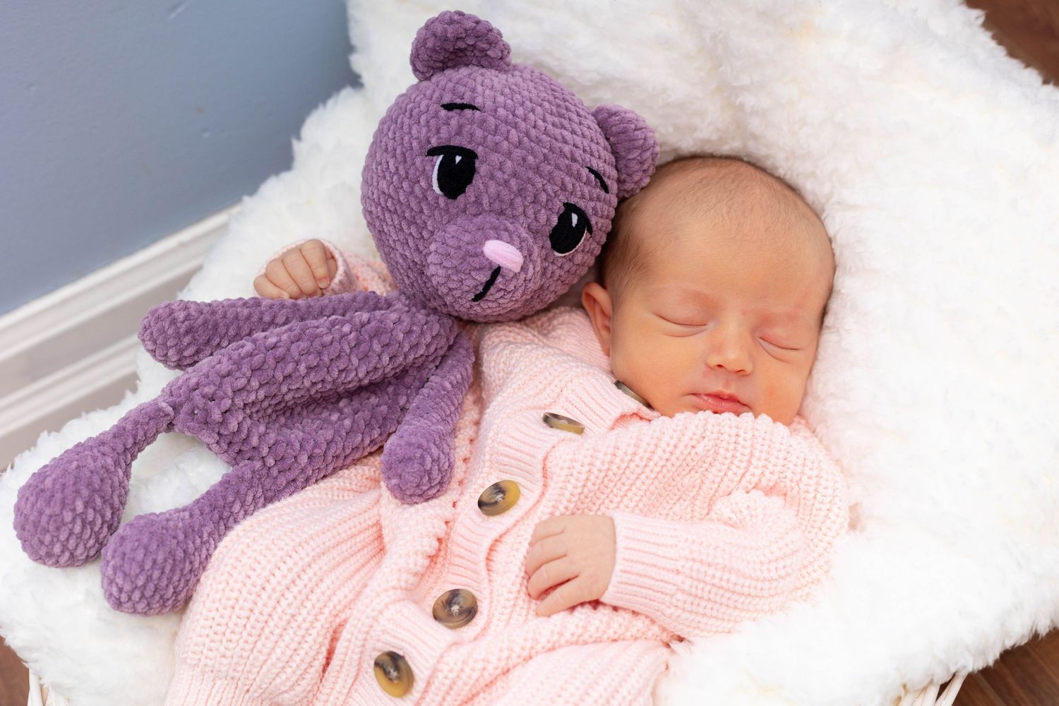 A sleeping newborn in a pink knit cardigan rests beside a purple teddy bear on a white fur blanket.