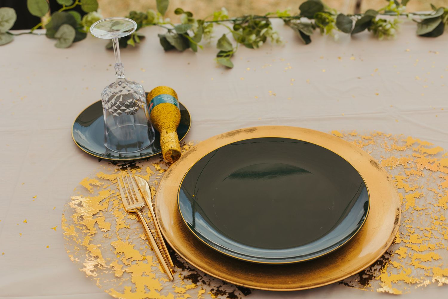 Elegant wedding table setting with black and gold plates, gold cutlery, and greenery centerpiece.