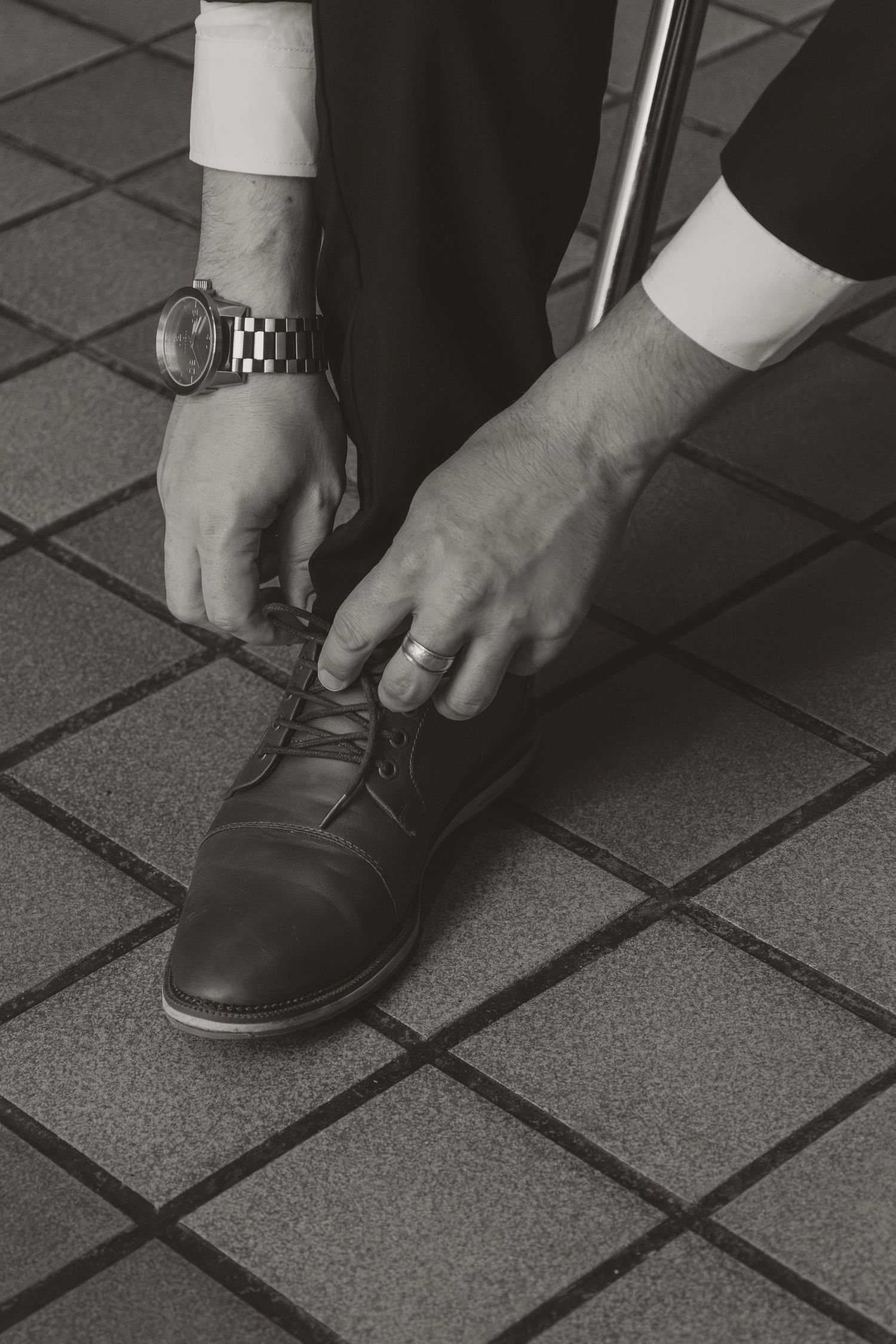 Groom tying dress shoe laces wearing a watch and wedding ring, black and white wedding detail.