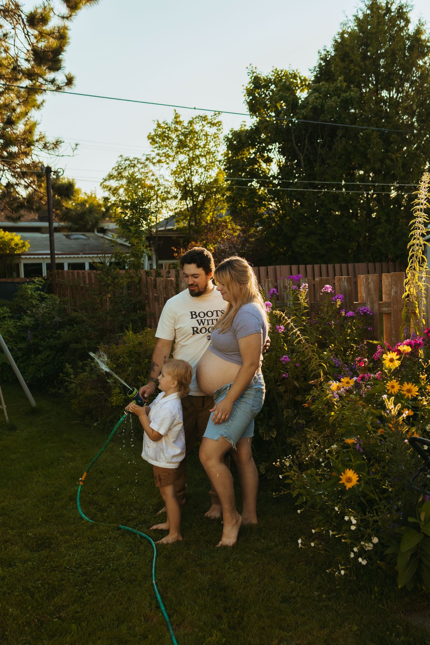Pregnant couple and young child playing with garden hose among colorful flowers in sunny backyard.