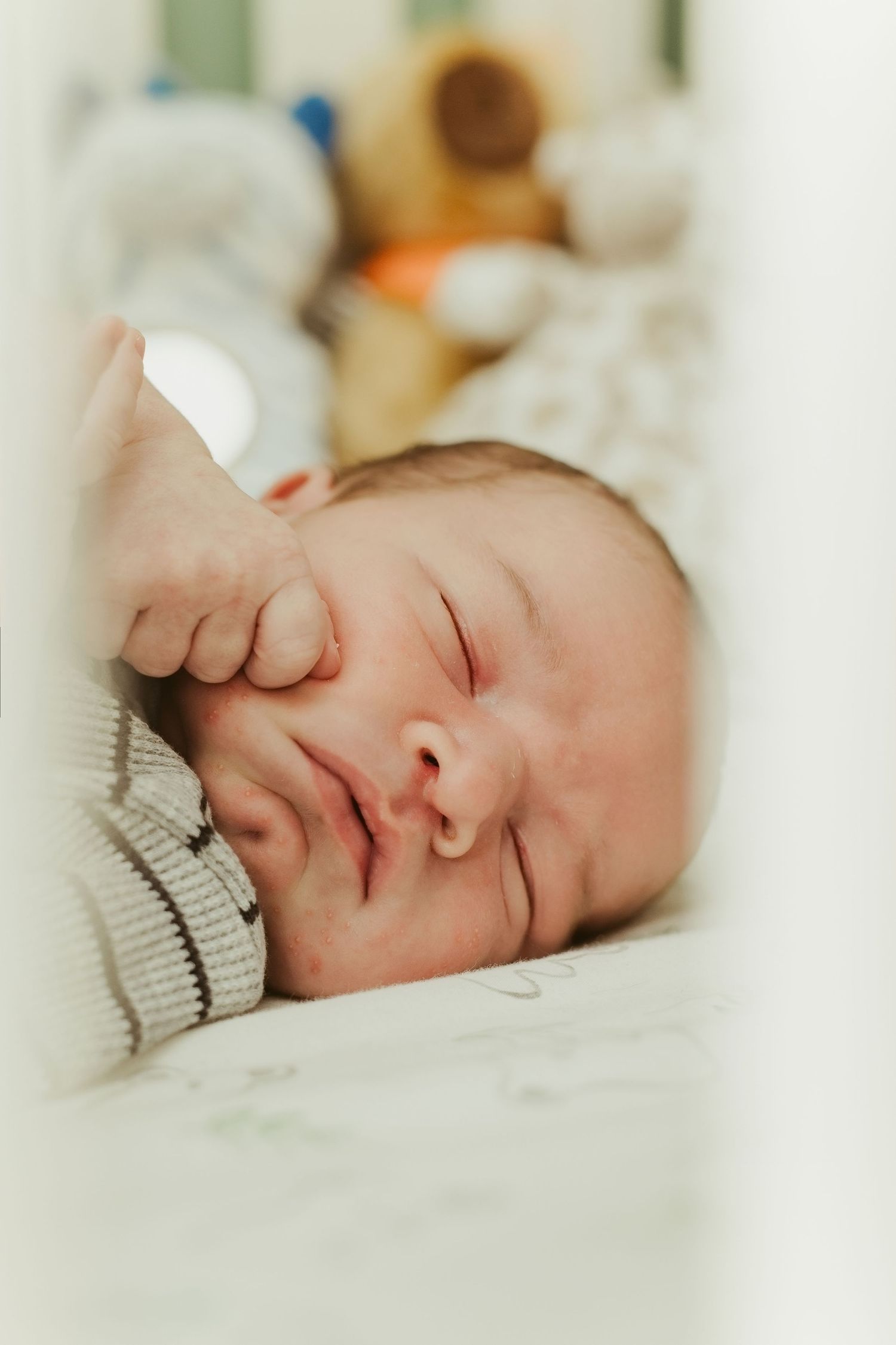 Peaceful newborn baby sleeping in a white crib with hand near face, stuffed animals in background.