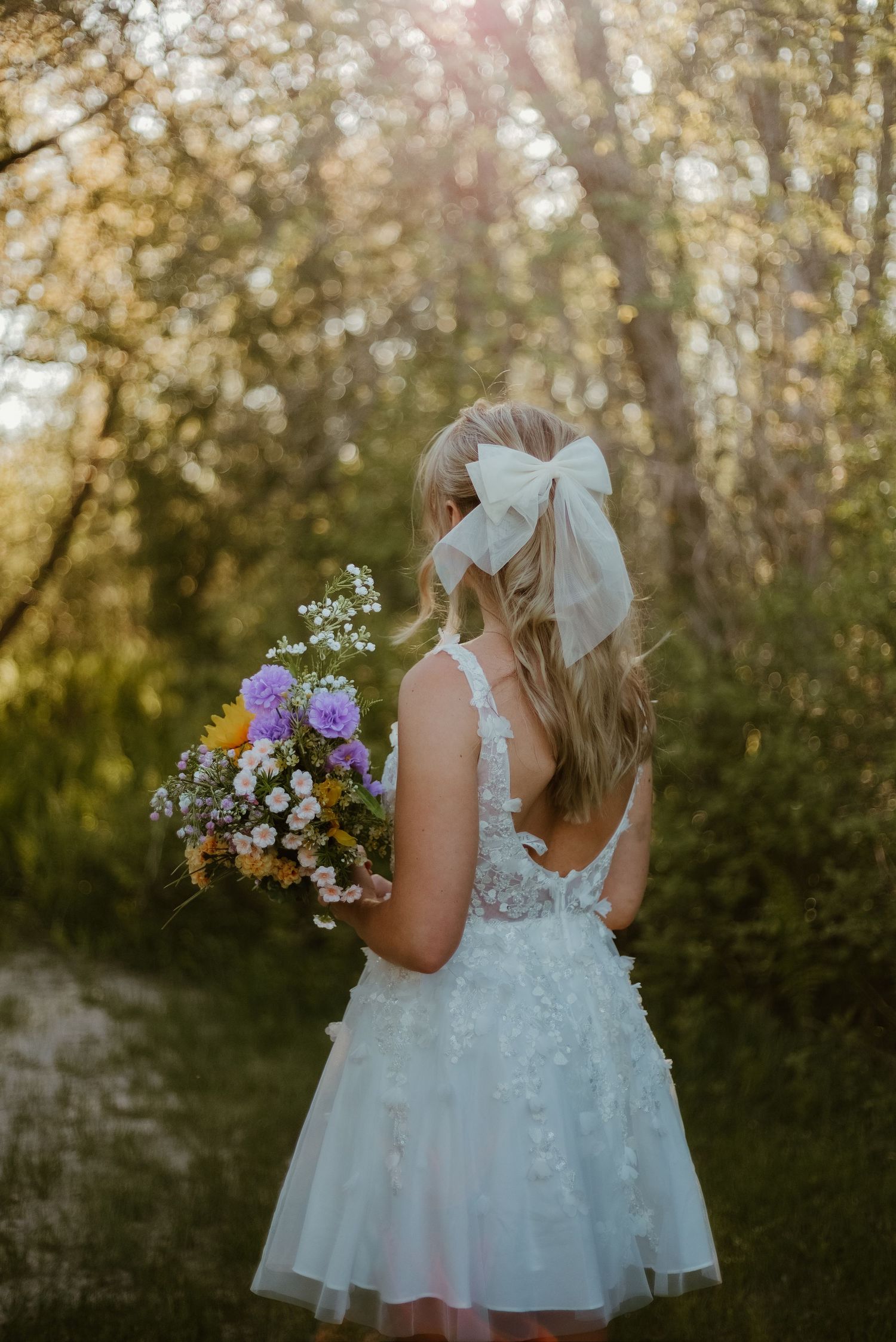 Bride in white lace dress holding wildflower bouquet, white bow in blonde hair, standing in sunlit forest path.