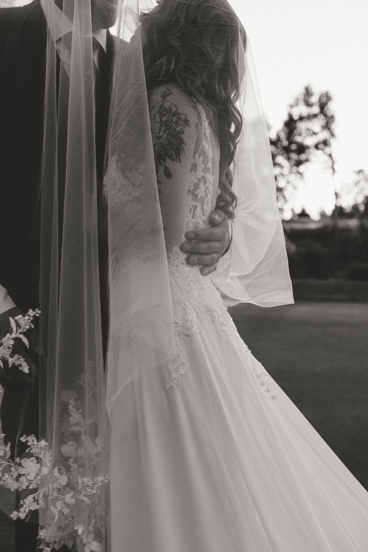 Black and white photo of bride in lace wedding dress with veil and floral back tattoo embracing groom outdoors.