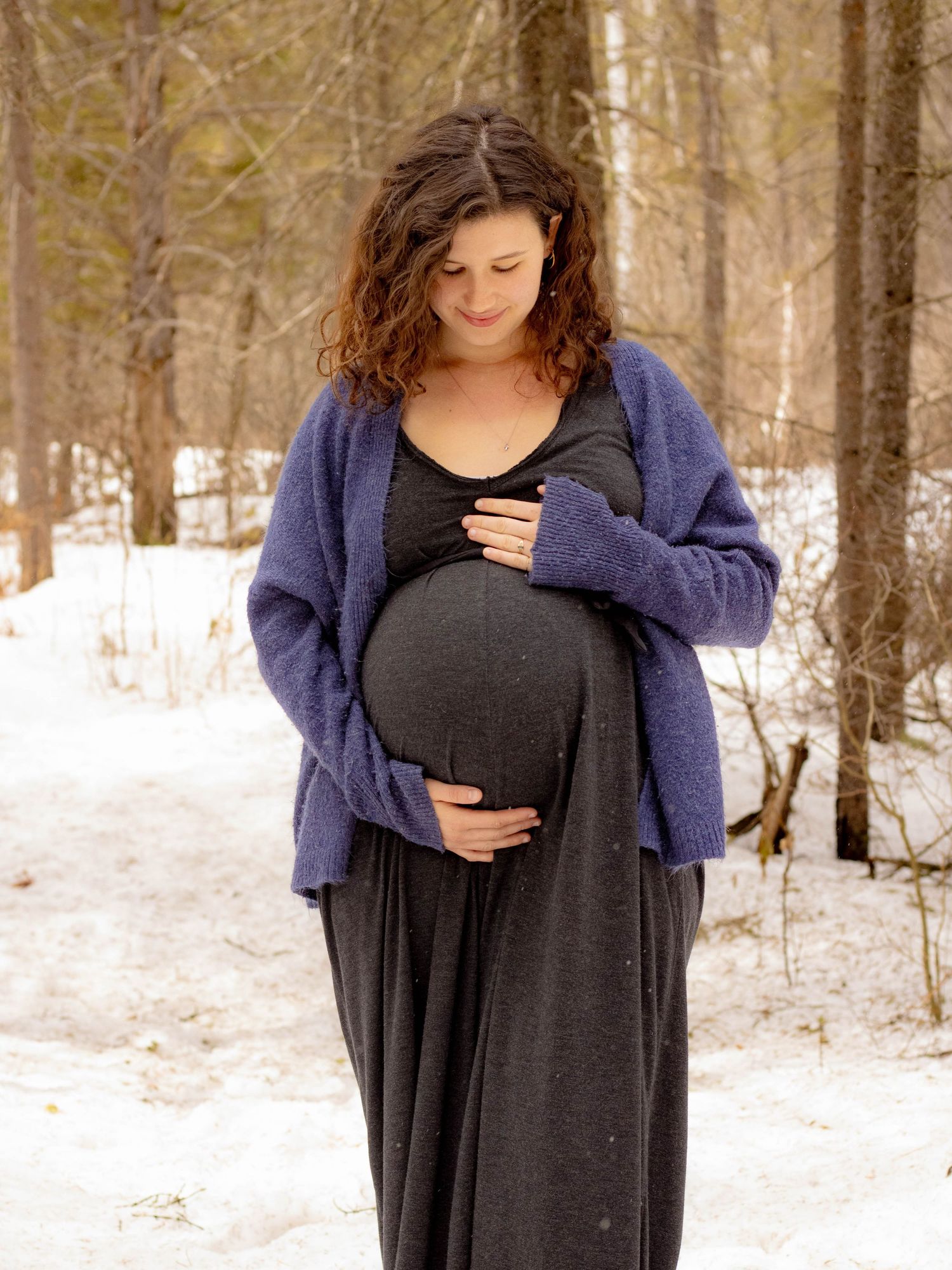 Pregnant woman in purple cardigan cradles baby bump in snowy winter forest setting.