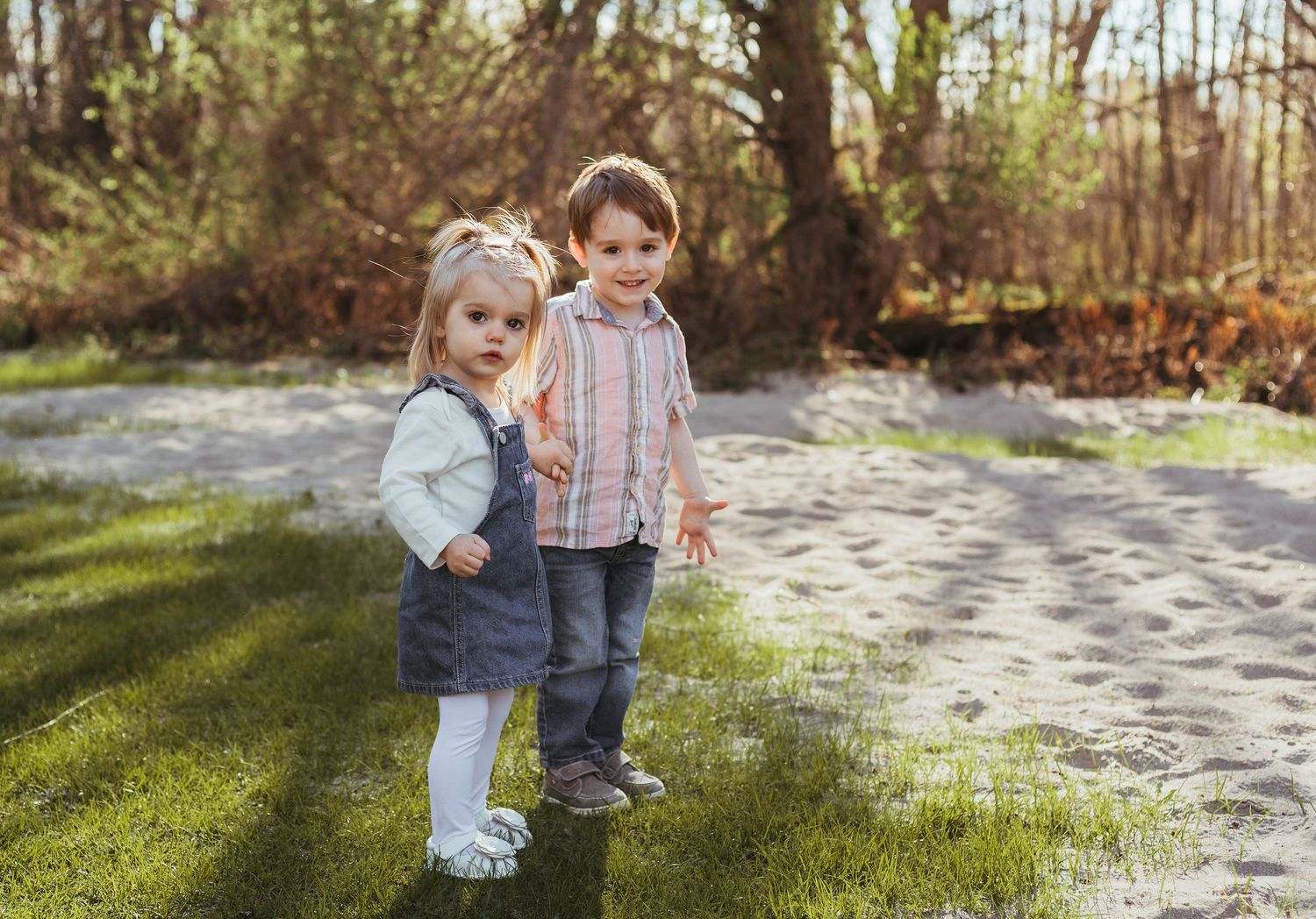 Two young toddlers, a girl in a denim dress and a boy in a plaid shirt, stand together outdoors on a sunny day.