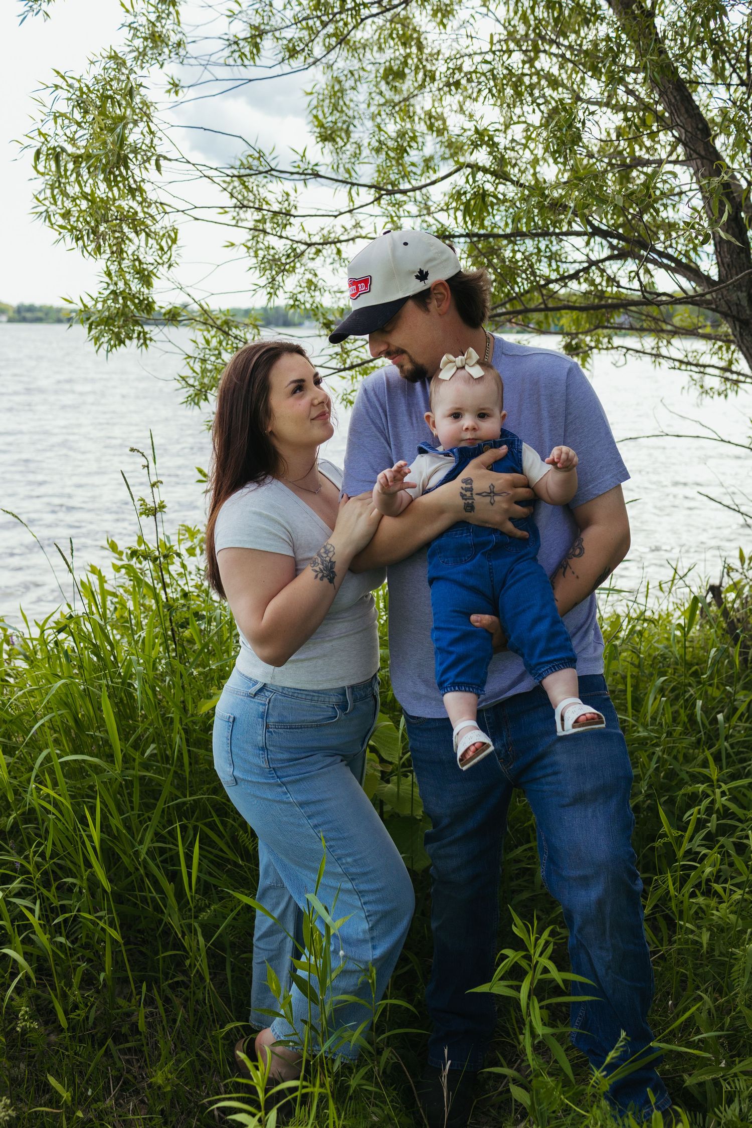 Young couple smiling at each other while holding their baby girl outdoors near a lake surrounded by lush green grass.