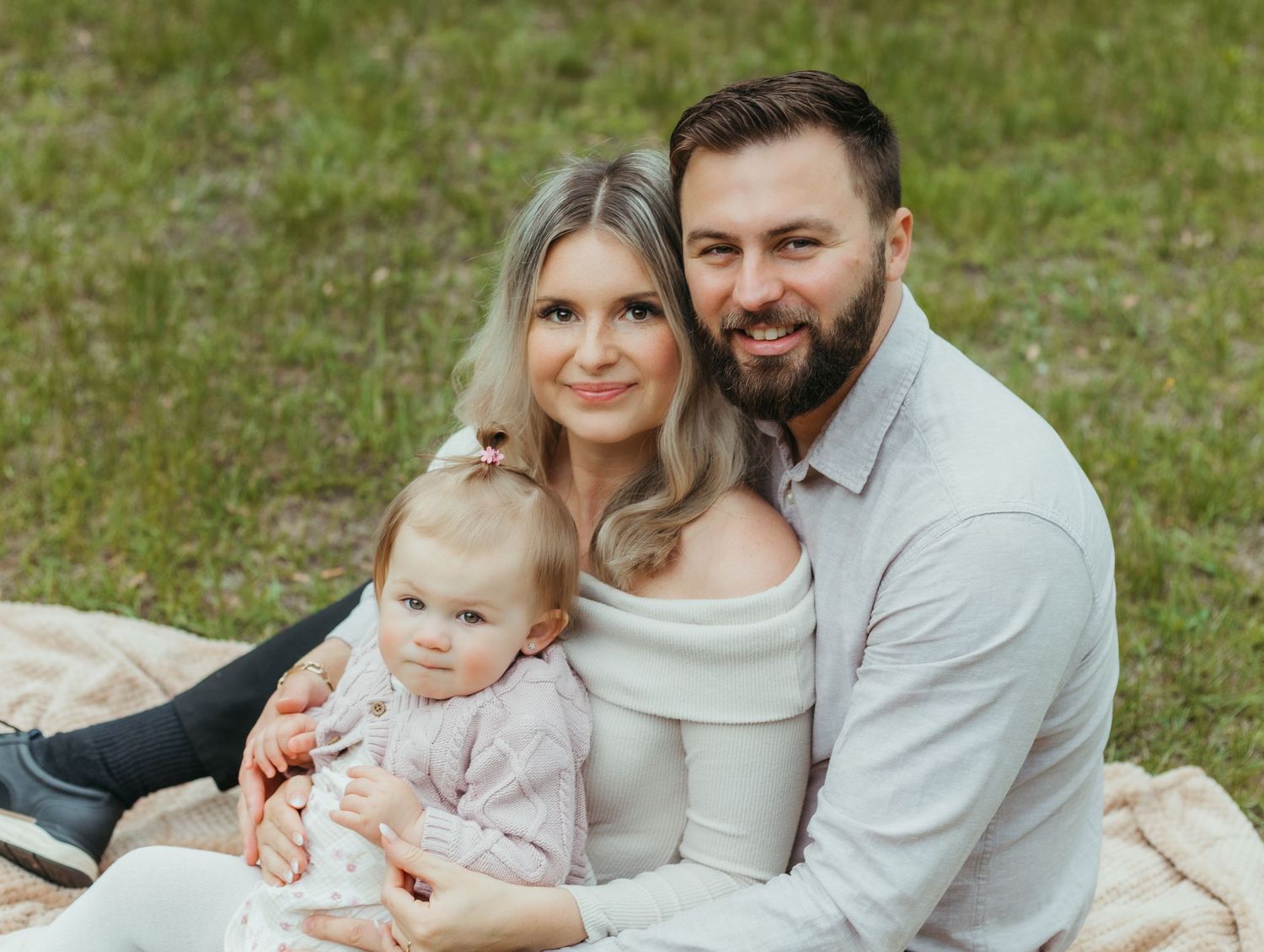 Young family with baby girl sitting on blanket in green field, smiling couple enjoying outdoor portrait session.