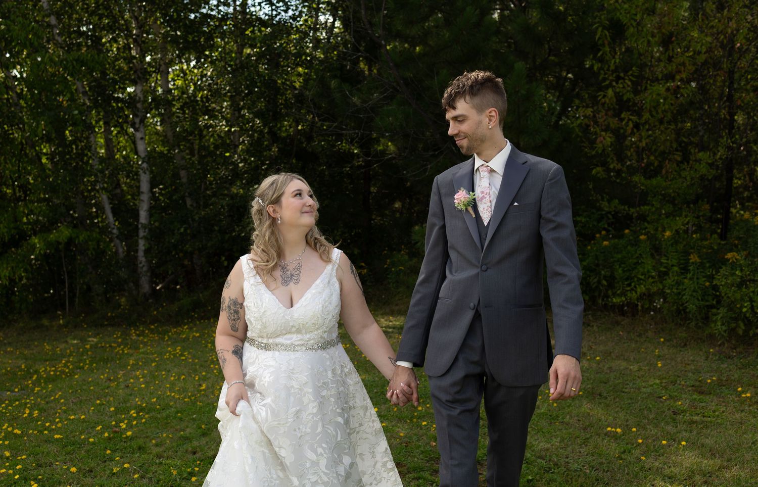 Bride and groom holding hands walking outdoors on green grass with trees in background on their wedding day.