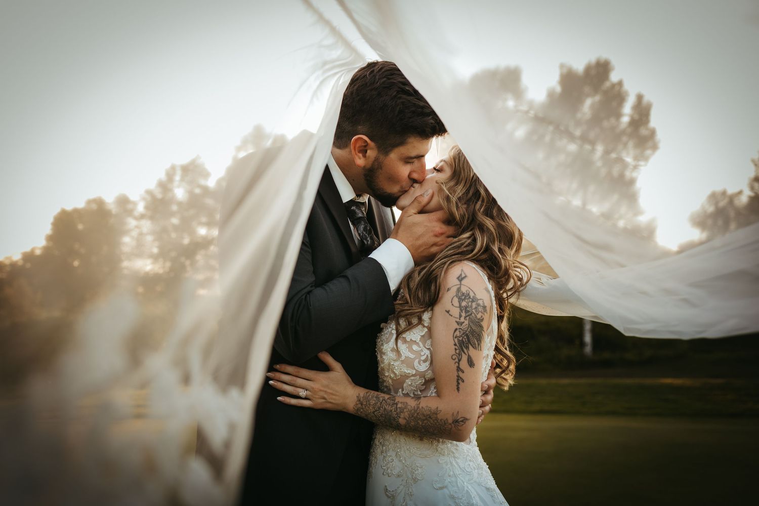 Bride and groom share a romantic kiss wrapped in a flowing veil at golden hour on a lush green wedding venue.