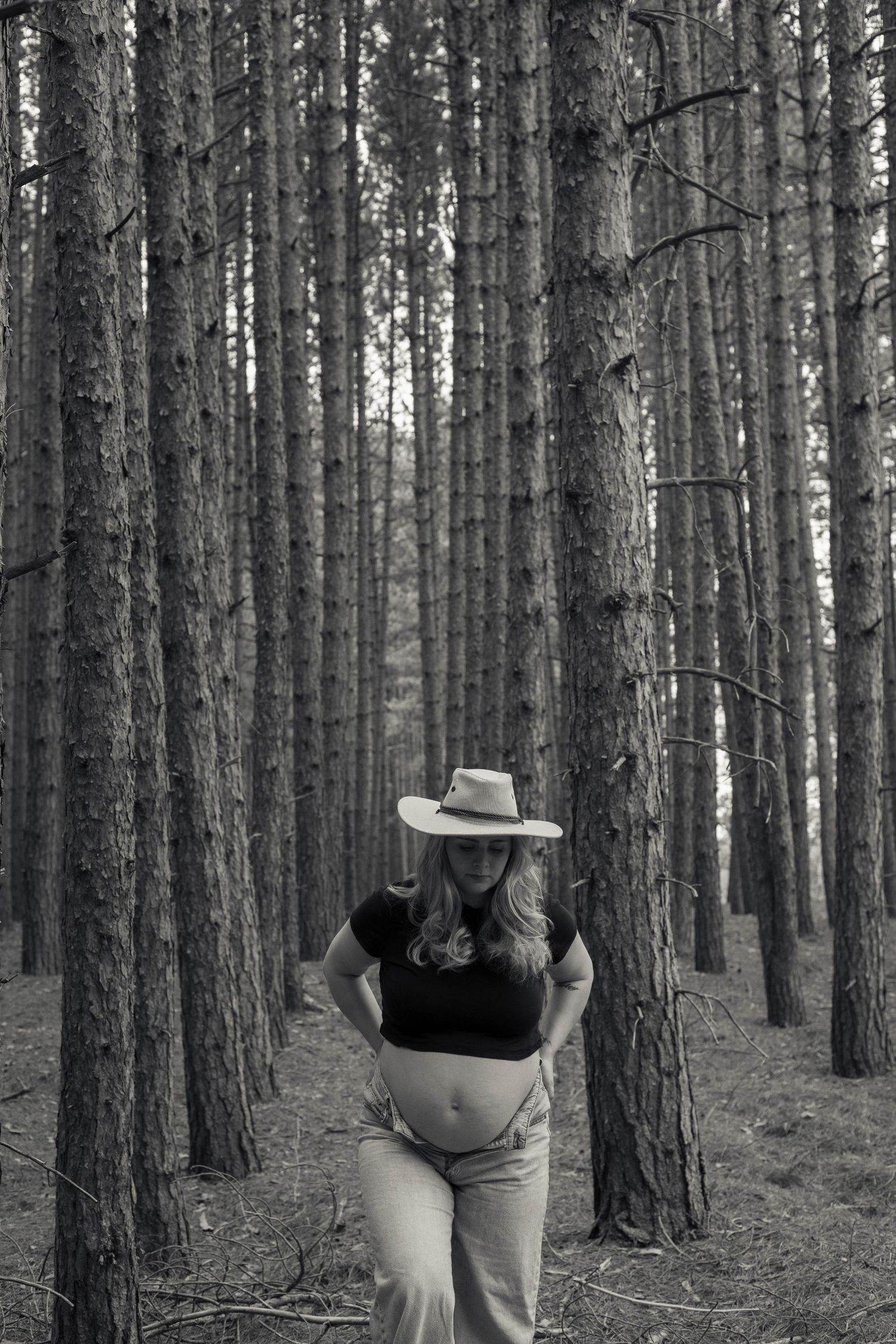 Black and white photo of a pregnant woman in a cowboy hat posing among tall pine trees in a forest.