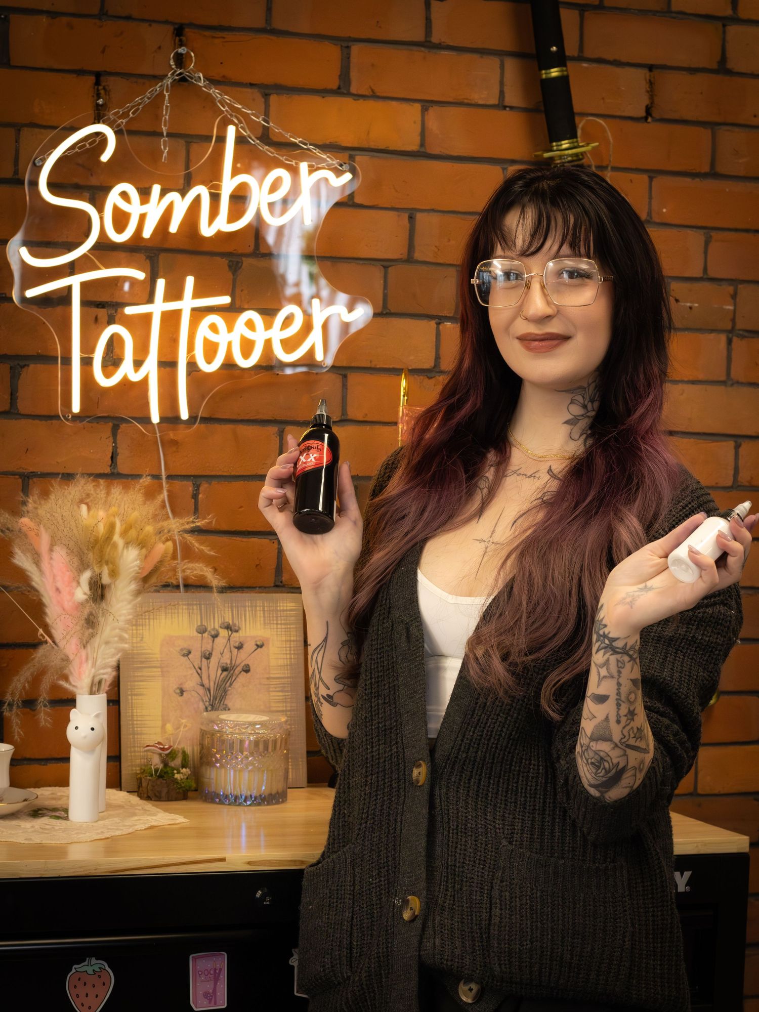 Female tattoo artist with purple hair holding ink bottles at Somber Tattooer studio.