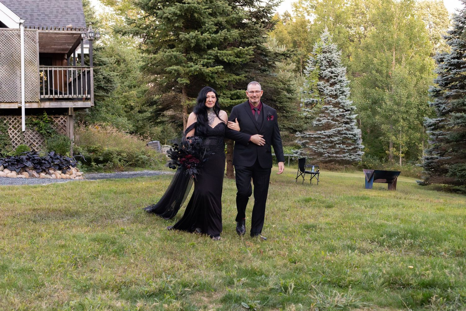 Gothic wedding couple in matching black outfits posing on green lawn with trees in background.