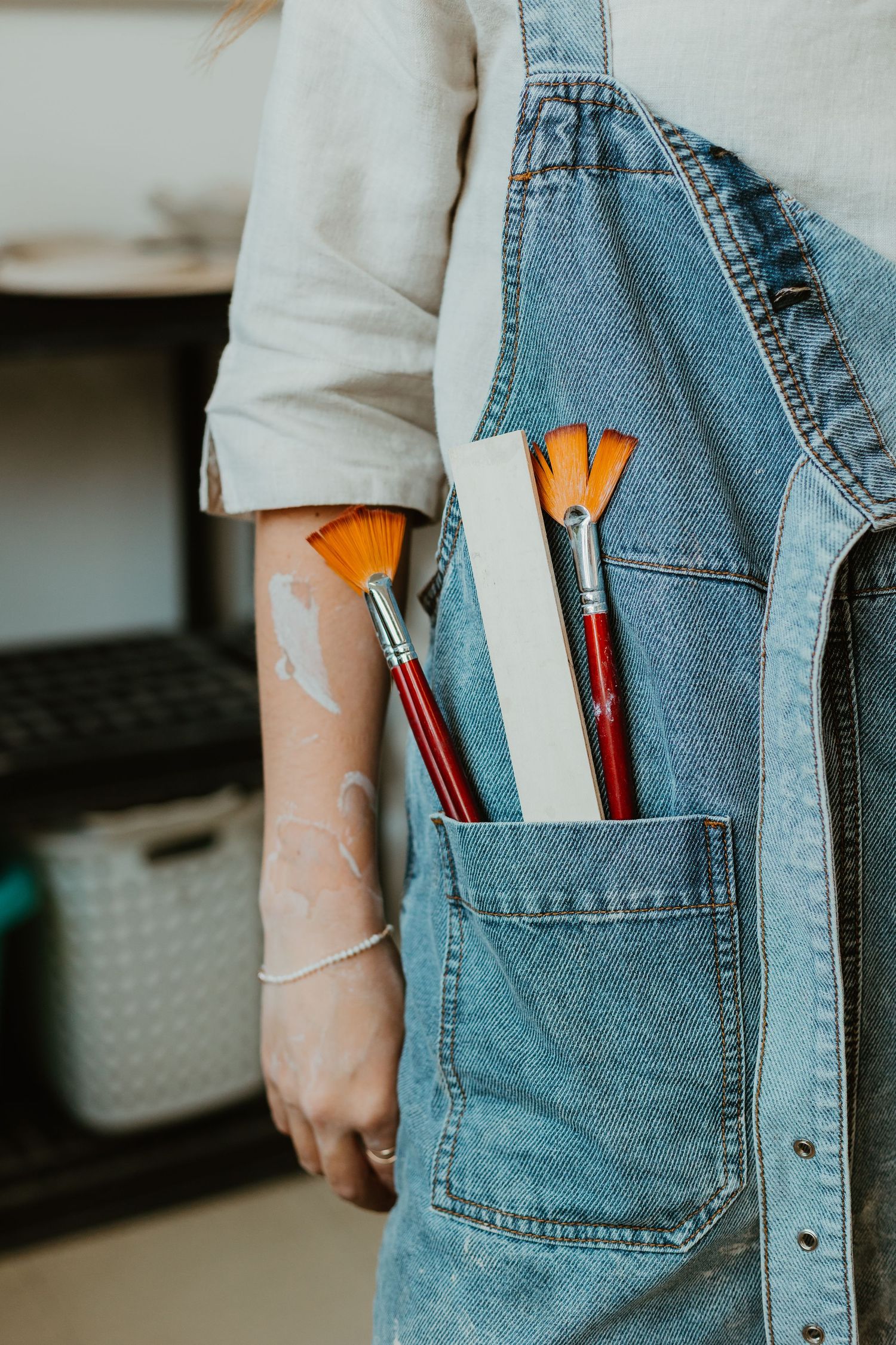 Artist in denim overalls holding paint brushes and a canvas in pocket, with paint on hand.