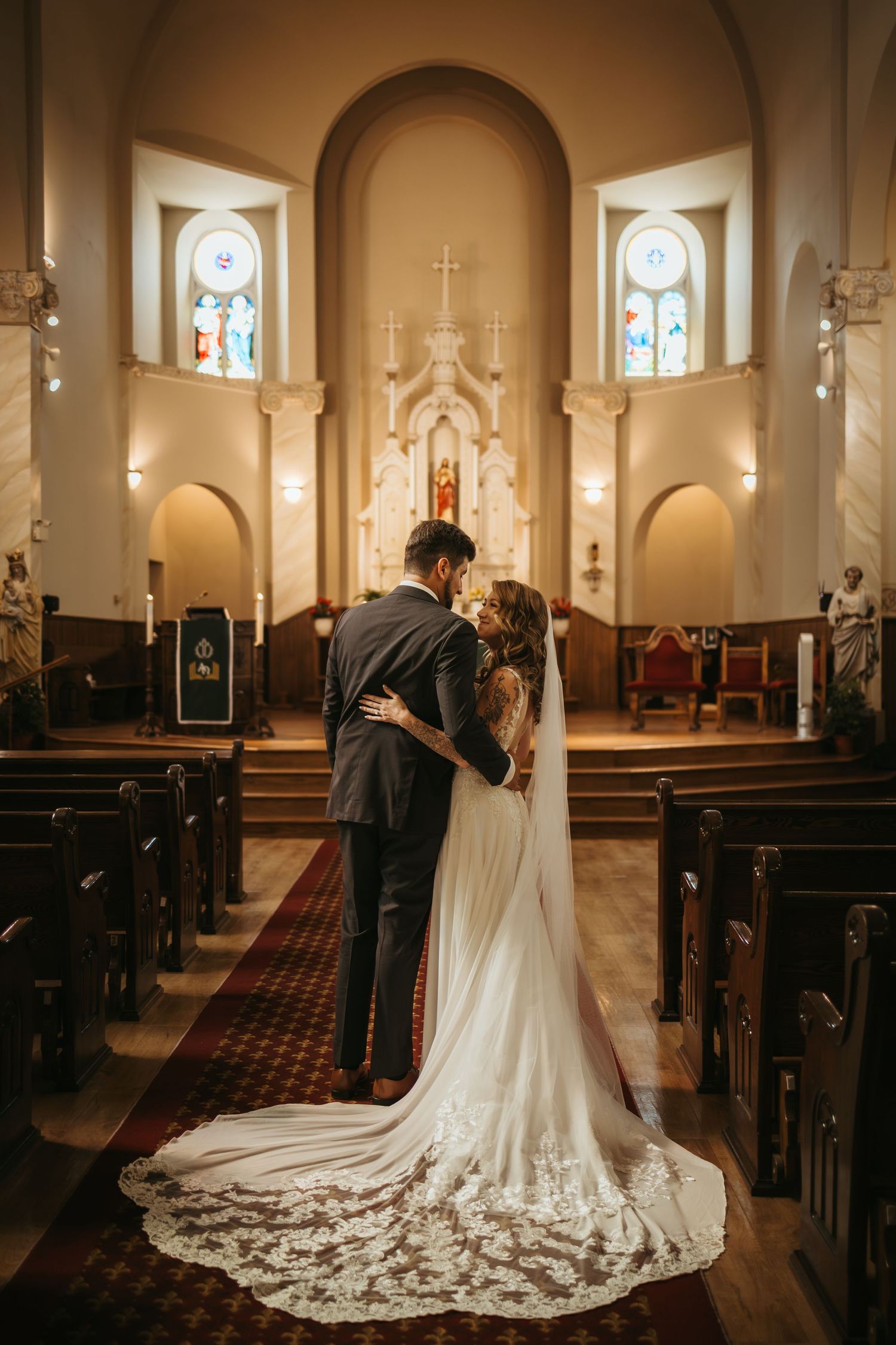Bride and groom share a kiss at the altar of an elegant church, with stained glass windows and a lace wedding gown train.