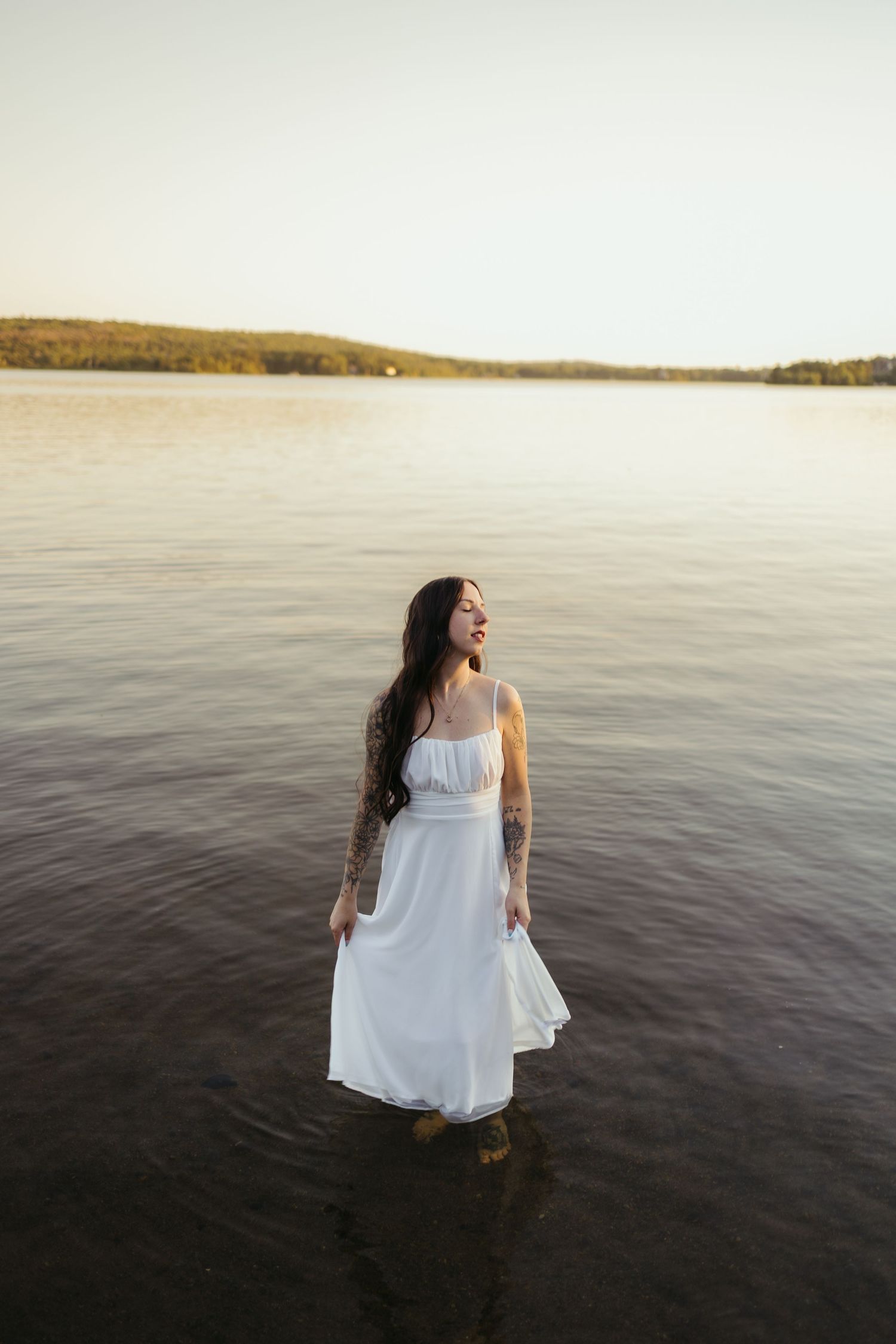 Woman in white dress standing in lake at sunset with tattoos and long dark hair.