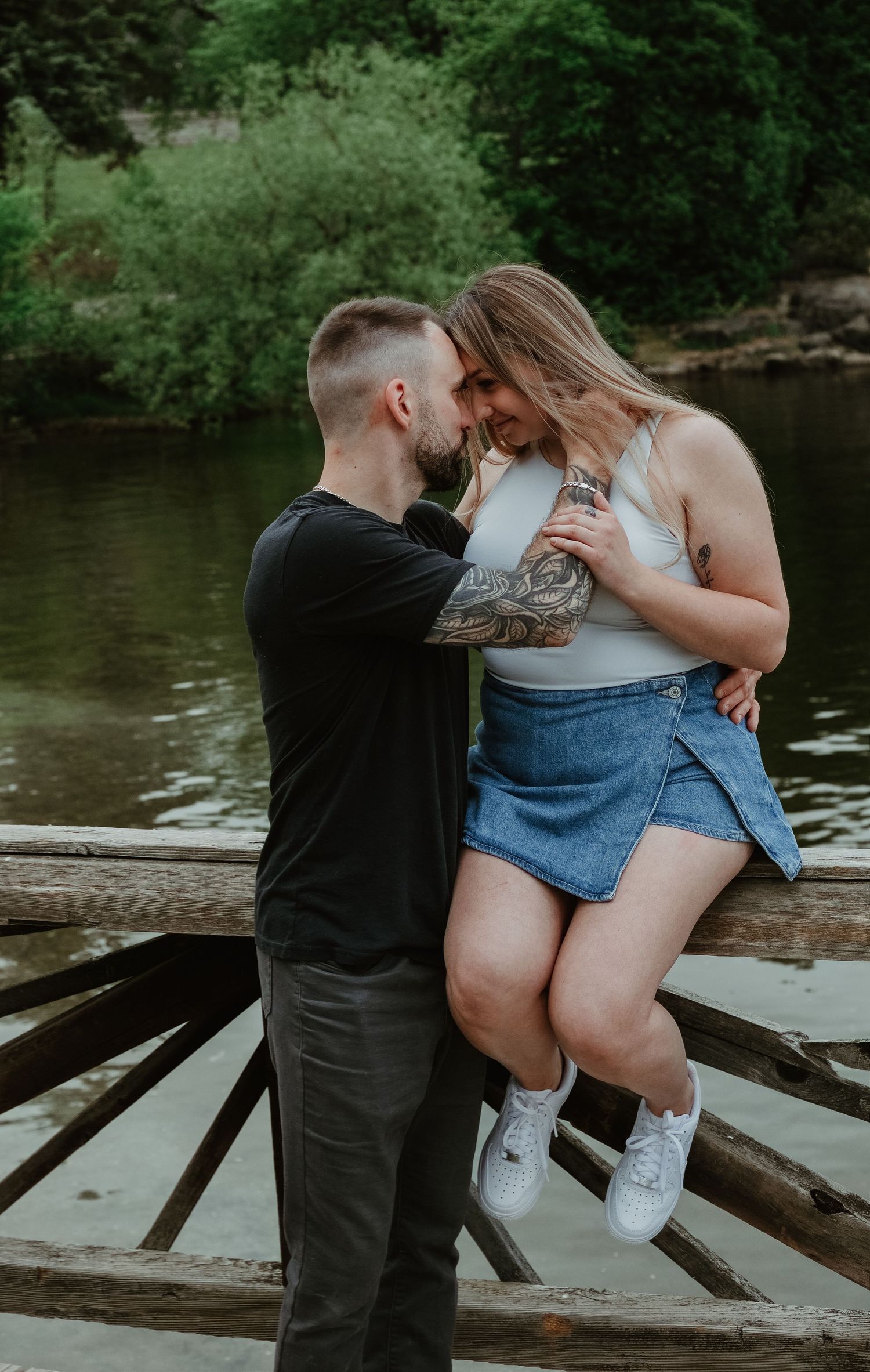 Couple sharing a romantic moment on a rustic wooden bridge by a lake, man with tattoos embracing woman in denim skirt.