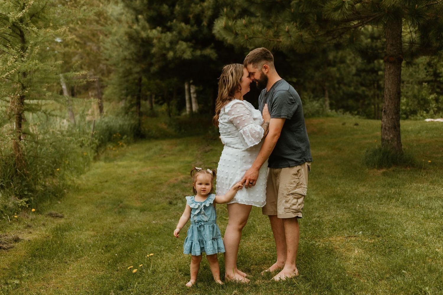 Family of three posing outdoors on green grass surrounded by pine trees, parents holding toddler girl in blue dress.