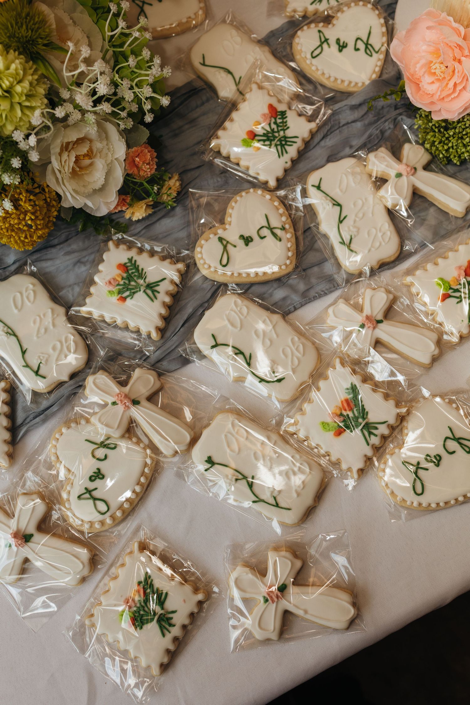Elegant white royal icing wedding cookies with green floral designs and initials, arranged on a table with flower bouquets.