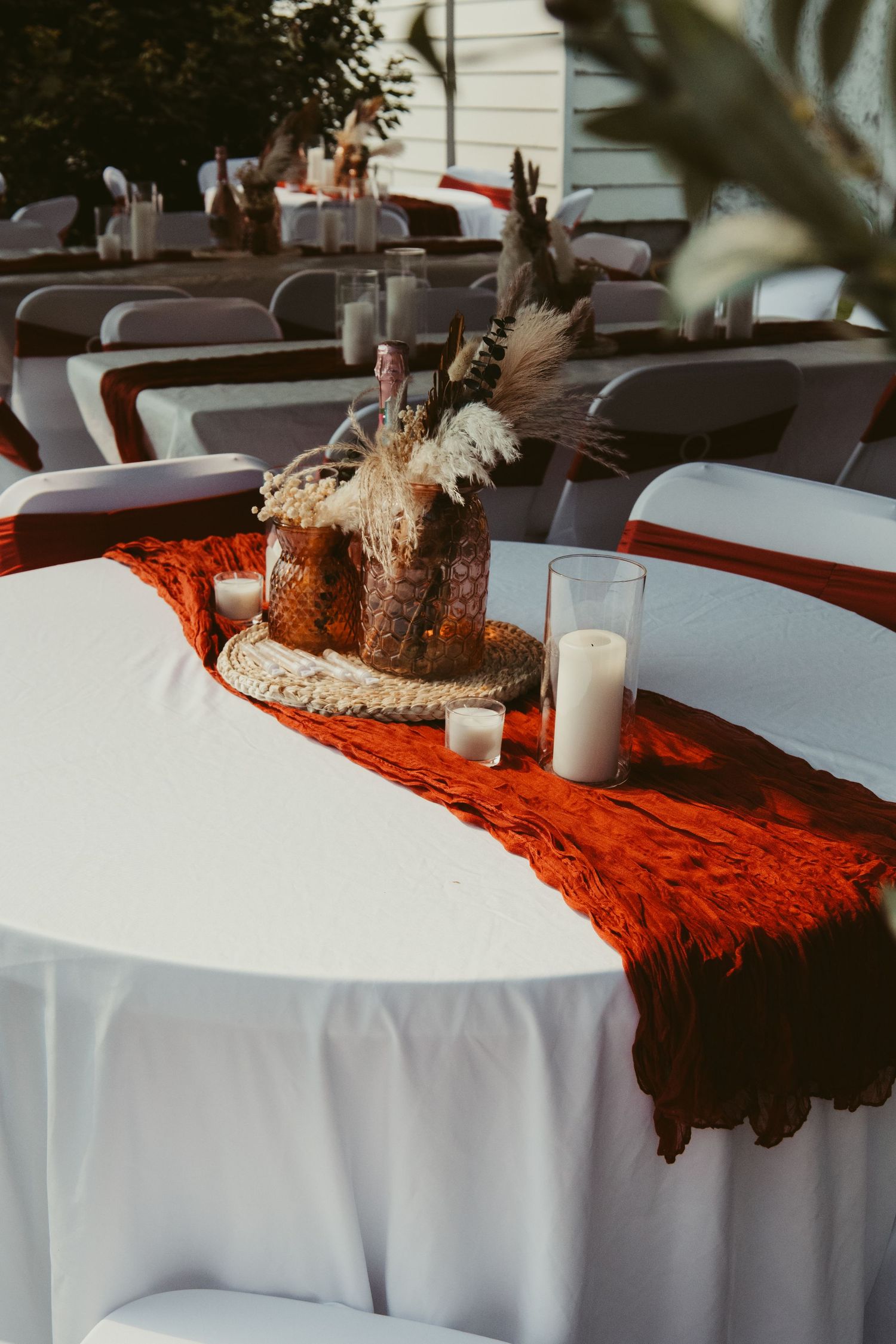 Elegant outdoor wedding reception tables with rust-colored runners, candles, and dried floral centerpieces.
