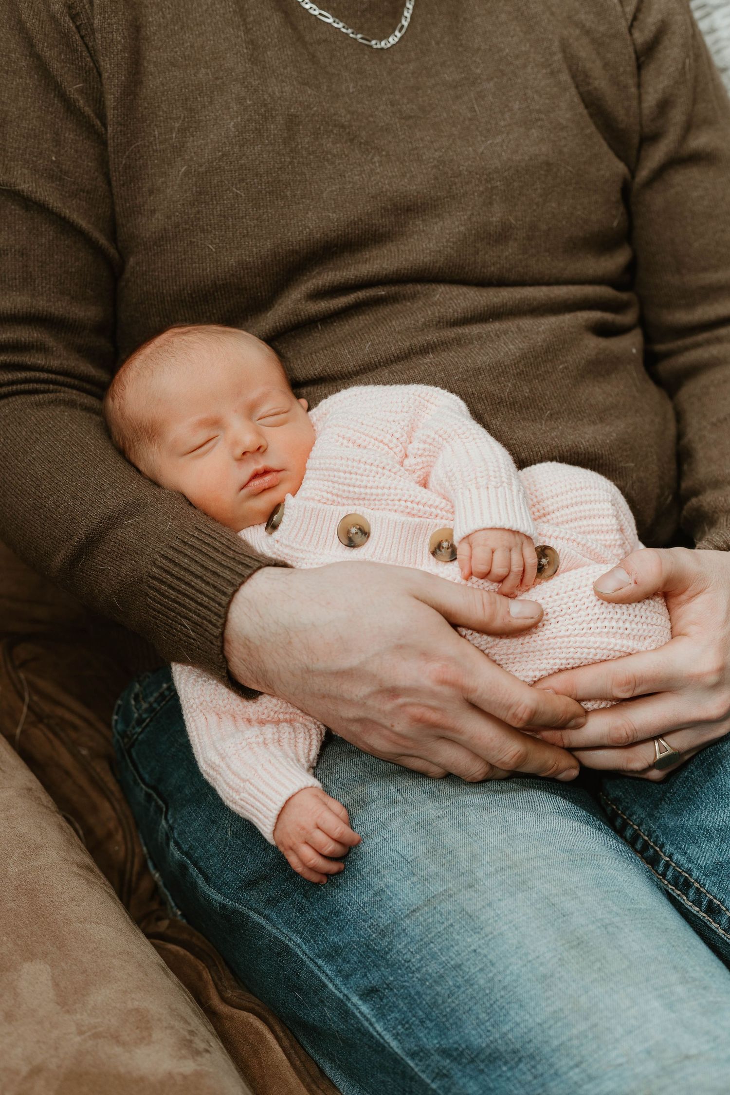 A newborn baby in a pink knit cardigan sleeps peacefully in a parent's arms on their lap.