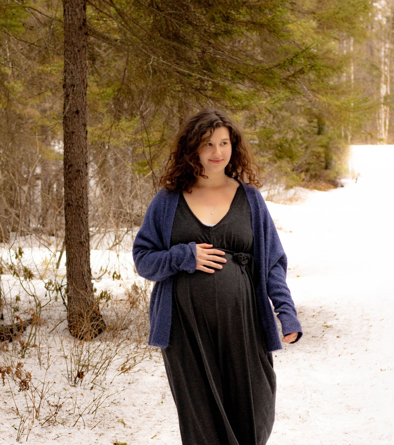Pregnant woman in black dress and blue cardigan smiling in a snowy winter forest setting.