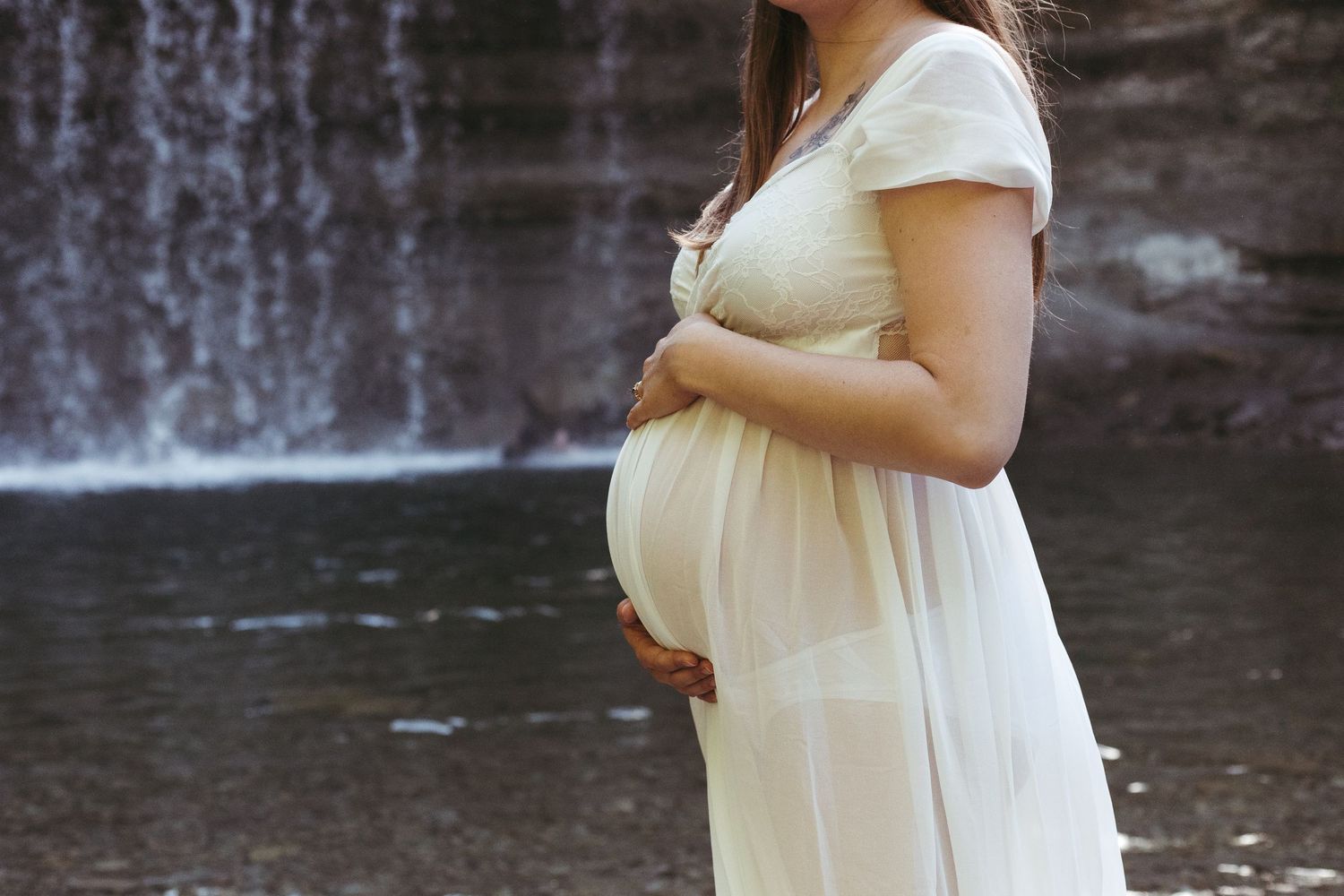 Side view of pregnant woman in white dress holding baby bump near a scenic waterfall.