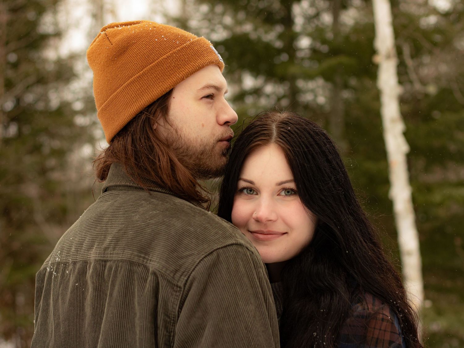 Couple embracing in snowy forest, man in orange beanie and green jacket with long-haired brunette woman smiling.