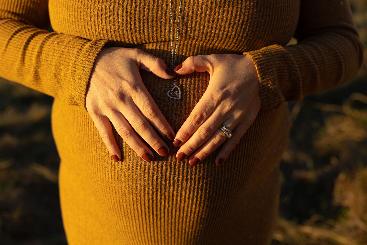 Close-up of pregnant woman in mustard dress forming a heart shape with her hands on her baby bump.