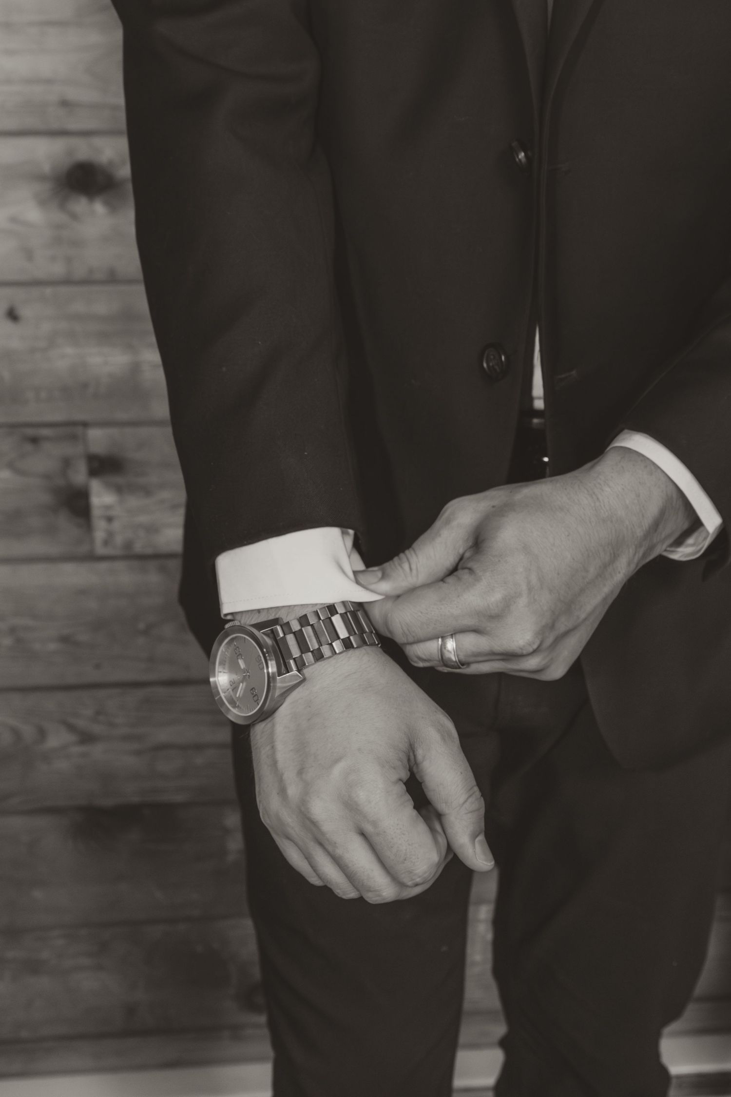 Groom in black suit adjusting white shirt cuff, wearing a watch and wedding ring, black and white.