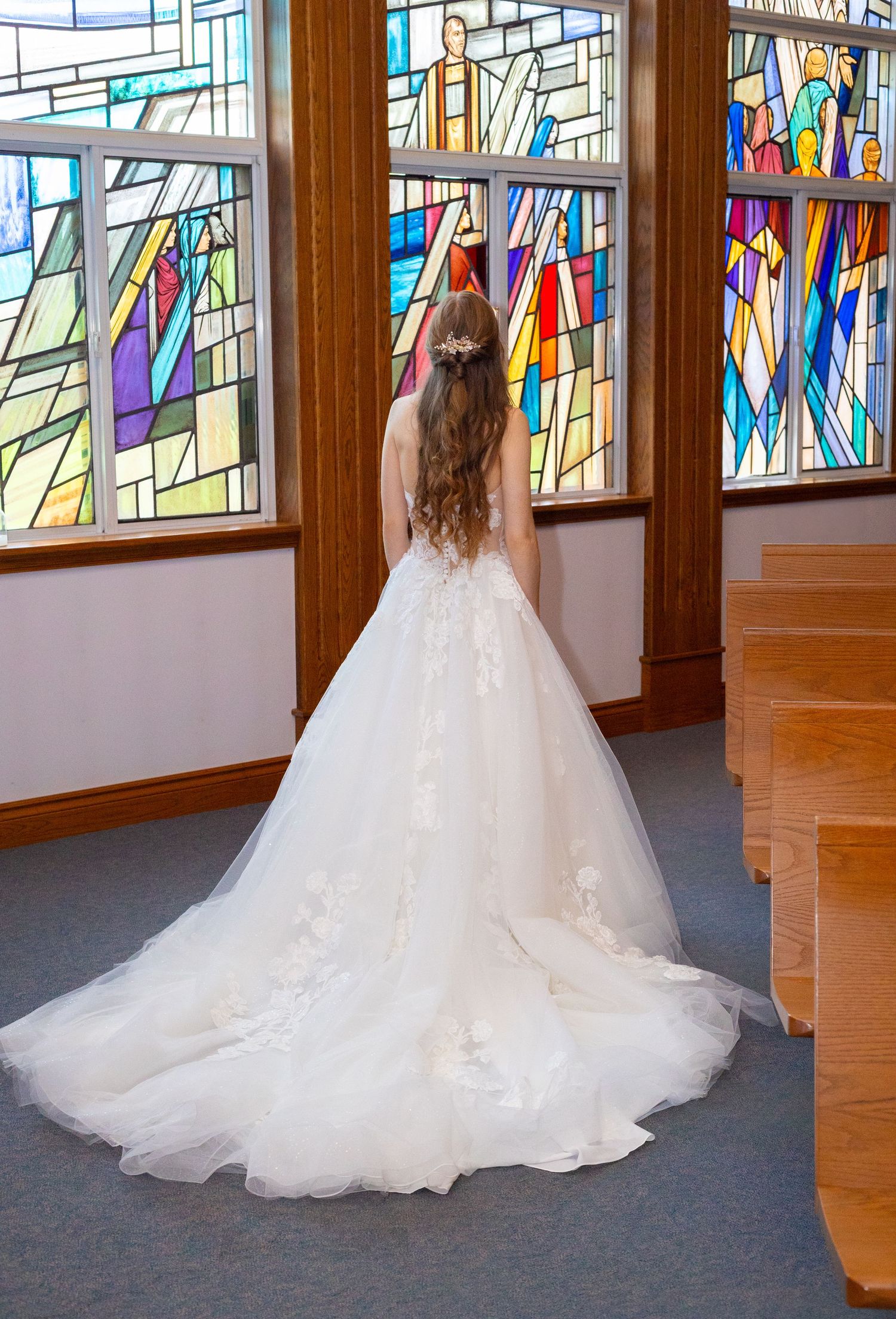 Bride in a stunning white ball gown with long train stands before vibrant stained glass church windows.