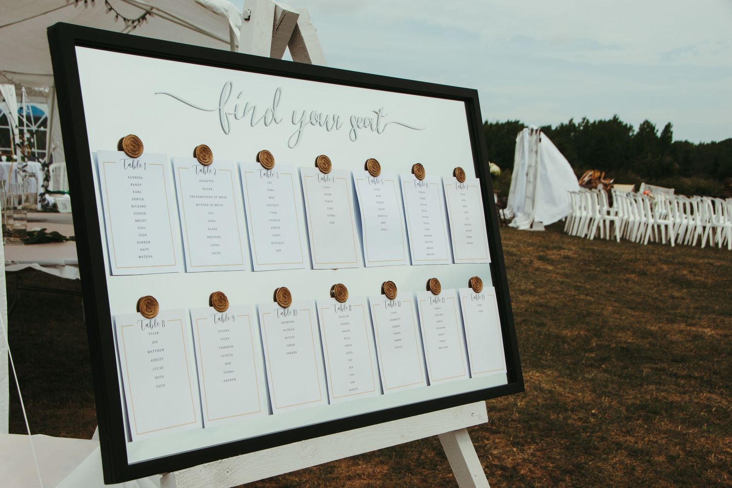Wedding seating chart with gold wax seals on easel at outdoor reception venue.