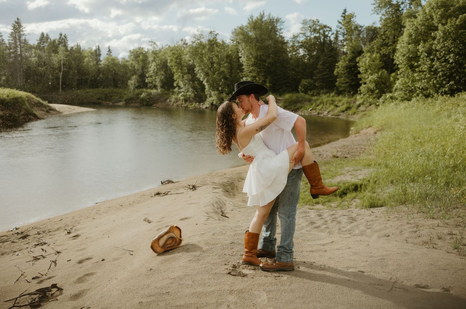 Couple sharing a romantic kiss on a sandy riverbank surrounded by lush green trees.