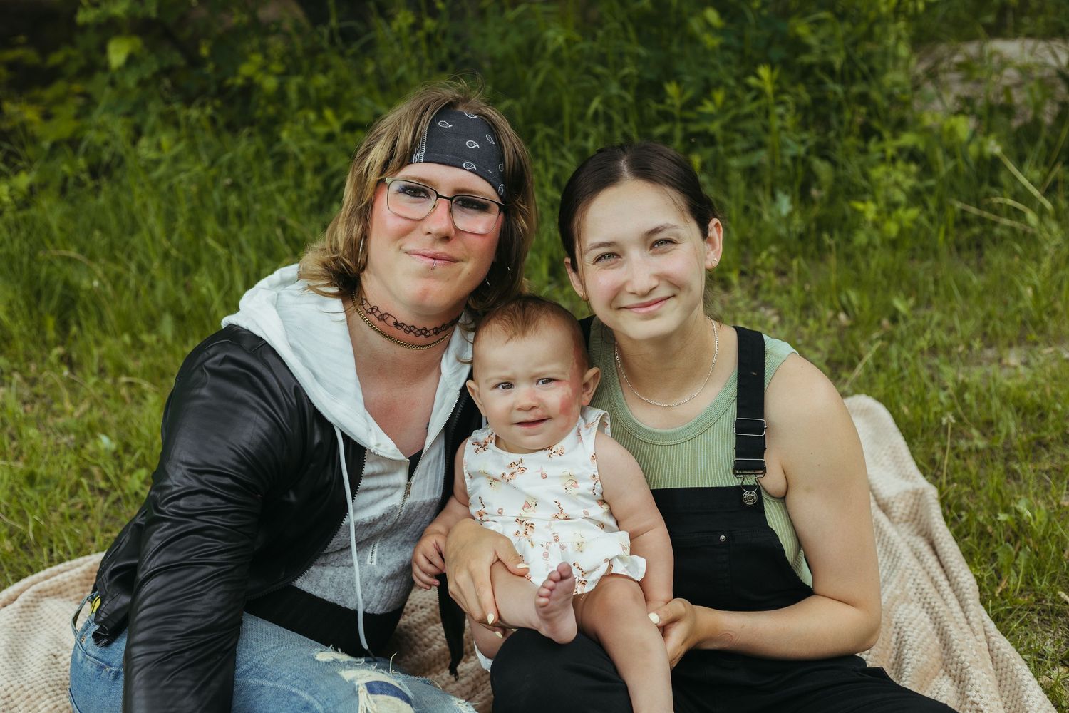 Two women and a baby sitting outdoors on a blanket, smiling together in a lush green nature setting.