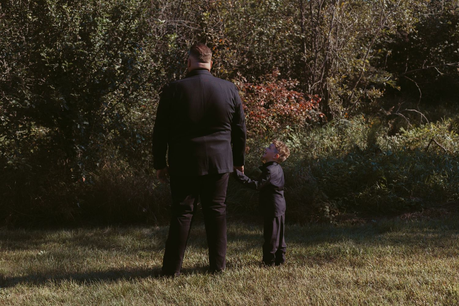 Man in dark suit holding hands with a young child in formal wear walking through autumn foliage.
