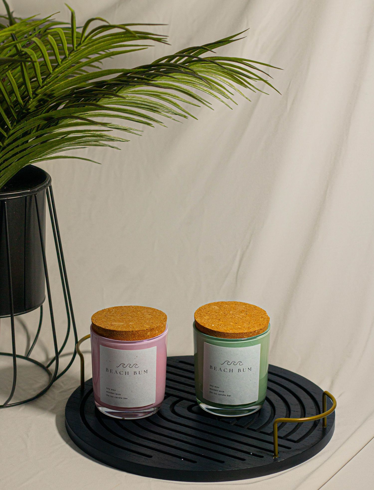 Two scented candles with cork lids in pink and green jars on a black tray beside a plant.