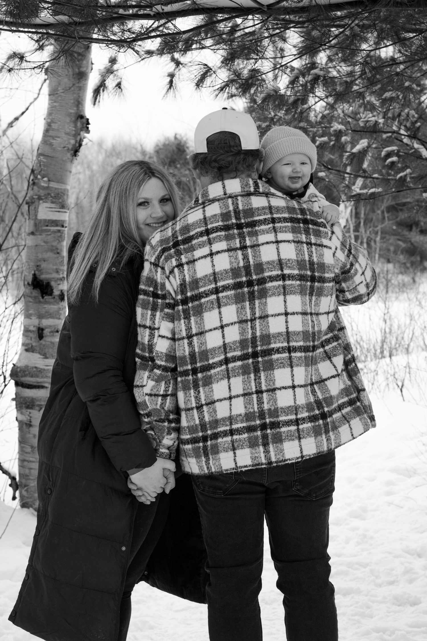 Black and white photo of a family in winter, father holding baby on shoulder, mother smiling beside him.