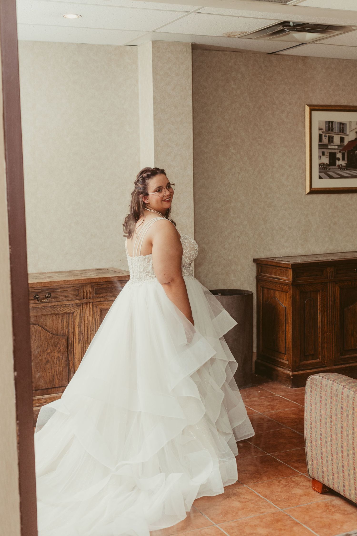 Bride in a layered white ball gown with lace bodice smiling over her shoulder in a hotel room.