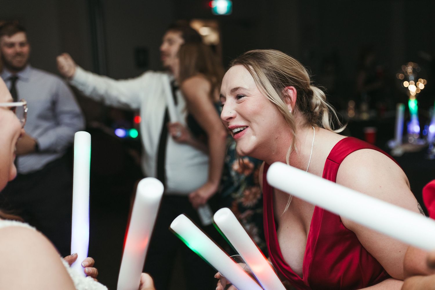 Woman in red dress laughing and dancing with glow sticks at a lively wedding reception party.
