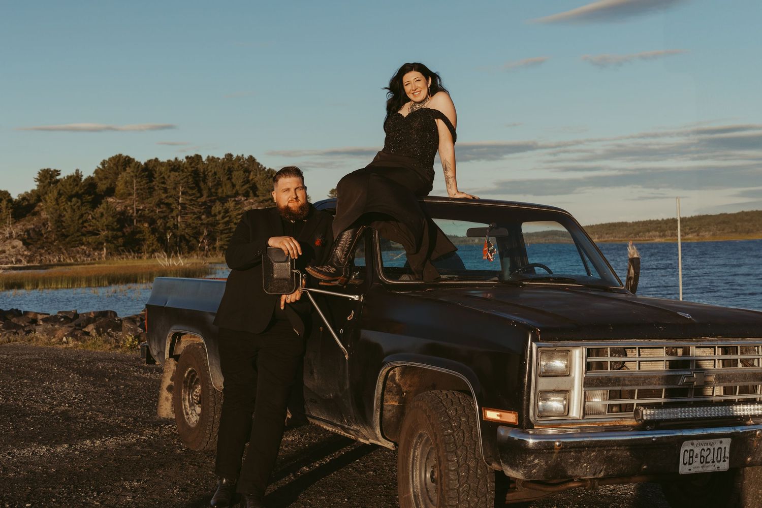 Laughing bride sitting atop a vintage black pickup truck with groom leaning against it by a lake.